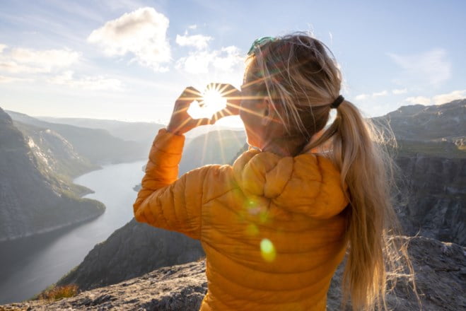 Jente står på toppen av et fjell og viser et hjerte med hendene mot sola.